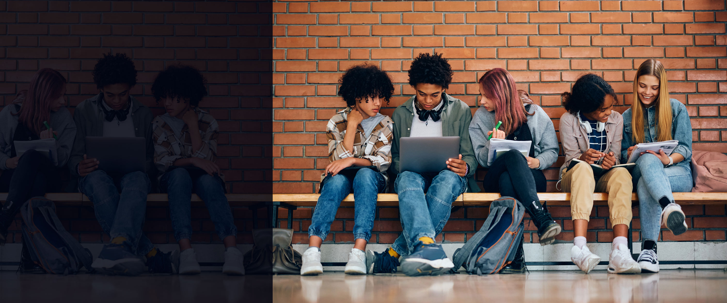 Students sitting together on a bench against a brick wall looking at laptops and homework